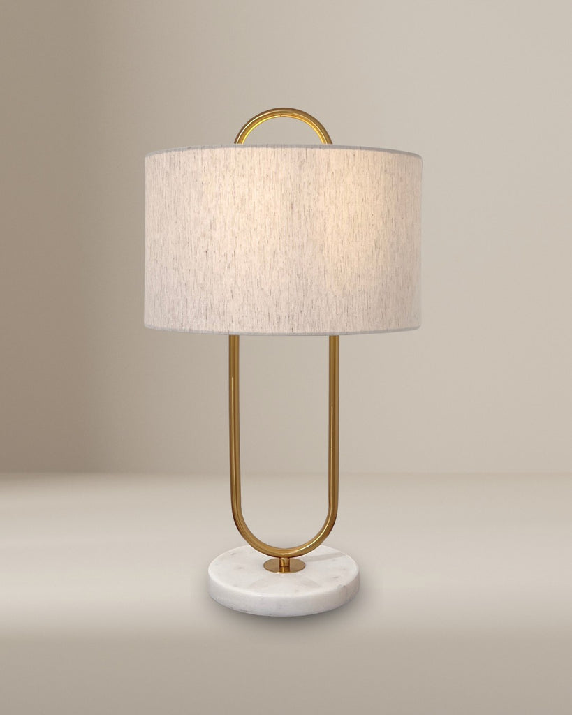 Minimalist table lamp with a brass U-shaped frame, white fabric shade, and circular marble base, photographed against a neutral beige background.