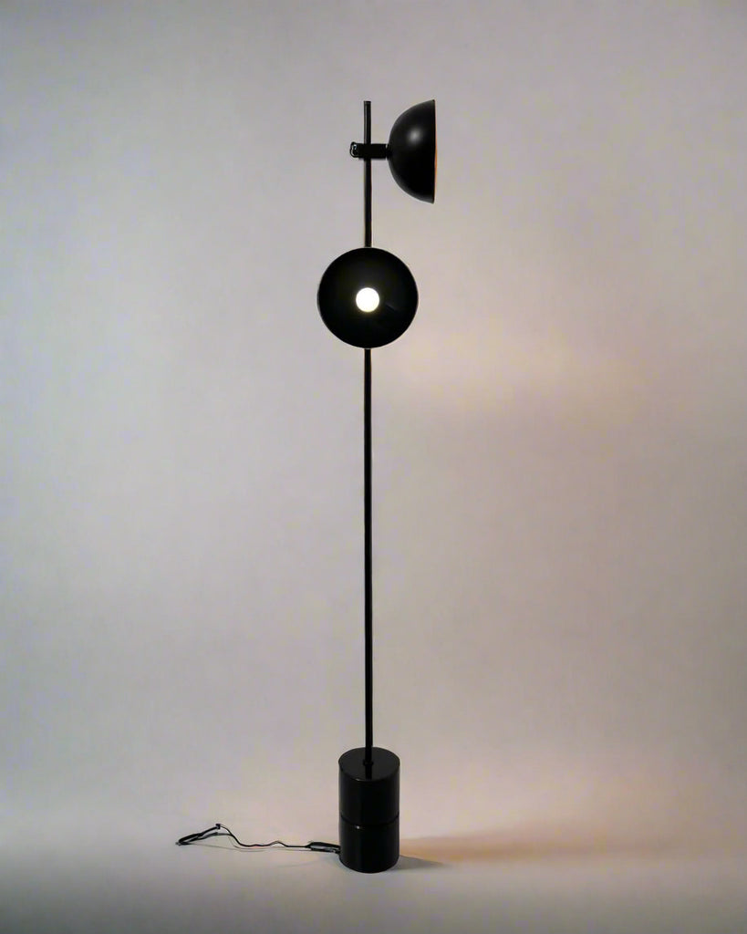 Minimalist black floor lamp with circular diffuser and top-mounted spotlight, standing on a cylindrical base against a neutral background.