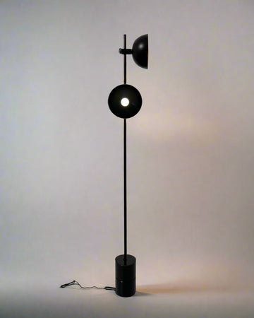 Minimalist black floor lamp with circular diffuser and top-mounted spotlight, standing on a cylindrical base against a neutral background.