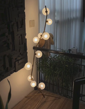 A softly glowing Feliz floor lamp with frosted glass globes and gold metal frame, illuminating a cozy interior with black textured wall art and indoor plants.
