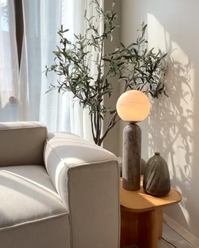 Baudet marble globe table lamp placed on wood side table beside ceramic vase and sofa, with olive branches casting shadows in natural sunlight