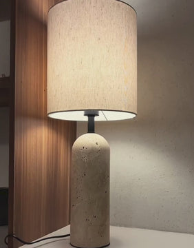 Kali Travertine Table Lamp placed against a wooden wall panel, showcasing its cylindrical natural stone base and soft ambient lighting ideal for cozy, refined interiors.
