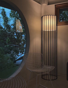 Video of a black Tuscan floor lamp with vertical slats creating a cozy nighttime glow in a modern room with circular window and white accent chair.