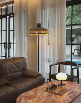 Video of rattan cane floor lamp with black iron base lighting up a cozy living room with leather sofa and sheer curtains.