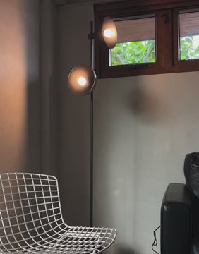 Black floor lamp with two lit bulbs in a room featuring a wireframe chair and wooden-framed window.