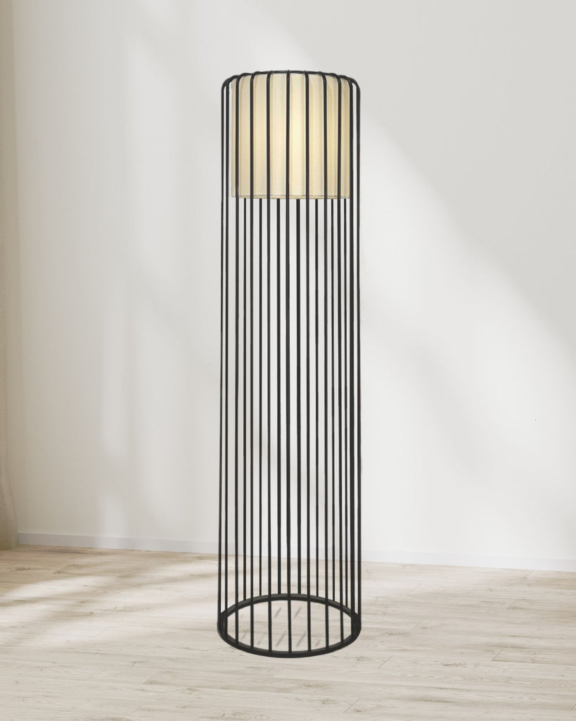 Tuscan-style floor lamp with a tall black wireframe cage design and warm inner light, standing on a light wood floor against a white wall.